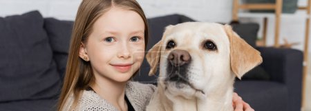 portrait of happy girl looking at camera while hugging labrador dog at home, banner