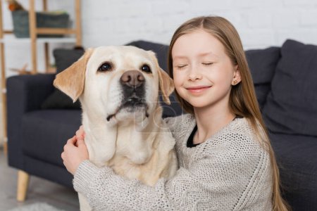 happy girl with closed eyes embracing labrador dog at home