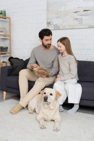 bearded man showing book to smiling daughter while sitting on couch near labrador dog