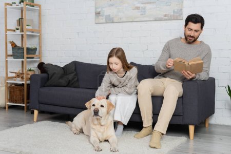 smiling bearded man reading book while sitting on couch near daughter petting labrador dog