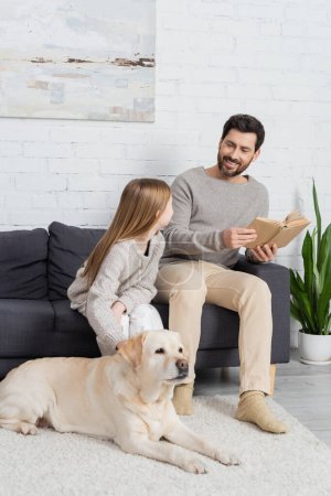 glücklicher bärtiger Mann sitzt mit Buch auf Couch und schaut Tochter neben Labrador-Hund im Wohnzimmer an