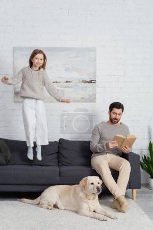 cheerful girl levitating above couch near dad with book and labrador dog on carpet