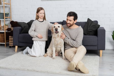 joyful father and daughter petting labrador dog on carpet in living room