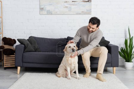 full length of happy bearded man sitting on couch in living room and petting labrador dog