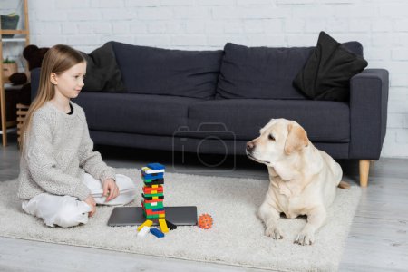 glückliches Mädchen und Labrador-Hund schauen einander in der Nähe von Holzklötzen Spiel und Laptop auf dem Boden im Wohnzimmer an