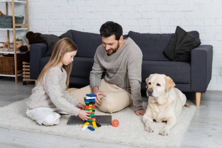 happy bearded man with preteen daughter playing wood blocks game near laptop and labrador dog on floor at home