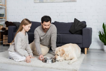 father and daughter sitting on floor carpet near bowl with water and labrador dog