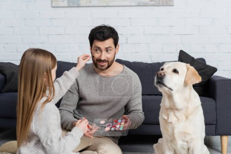 preteen girl holding makeup palette while playing with smiling dad near labrador dog in living room