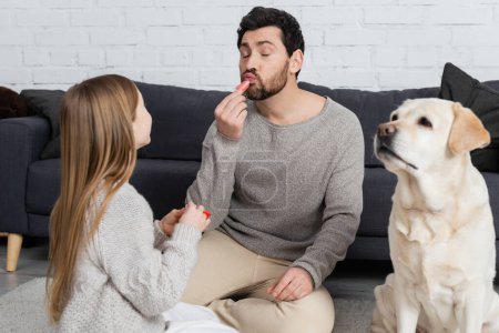 bärtiger Mann mit geschlossenen Augen, der Lipgloss aufträgt, während er mit Tochter in der Nähe von Labrador-Hund im Wohnzimmer spielt