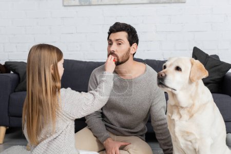 Frühchen besprüht Vater beim Spielen mit Labrador-Hund im Wohnzimmer mit Lipgloss