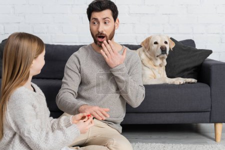 girl wearing ring on finger of amazed father while playing near labrador dog lying on couch in living room