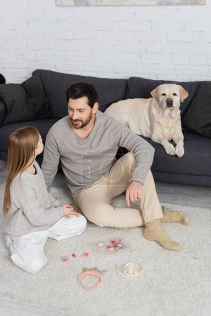 Labrador dog lying on couch near father and daughter sitting on floor near toy crowns and makeup palette 