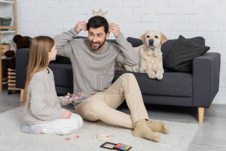 cheerful bearded man wearing toy crown while sitting on floor near daughter and dog lying on sofa