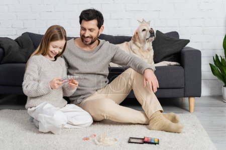 happy father and daughter looking at makeup palette while sitting near labrador dog in living room 
