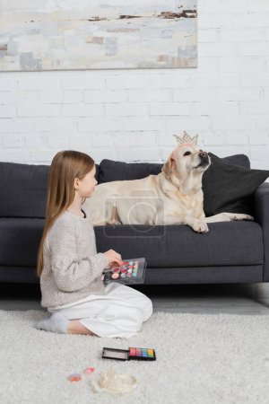 happy preteen girl holding makeup palette and looking at labrador in toy crown sitting on couch 