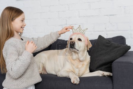 happy preteen girl adjusting toy crown on labrador lying on couch 
