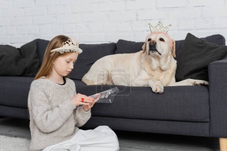 preteen girl in crown looking at makeup palette while sitting near labrador on couch 