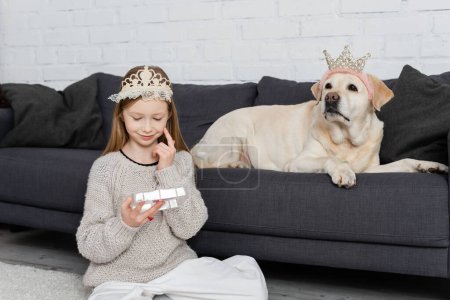 happy preteen girl in crown looking at makeup palette and sitting on carpet near labrador 