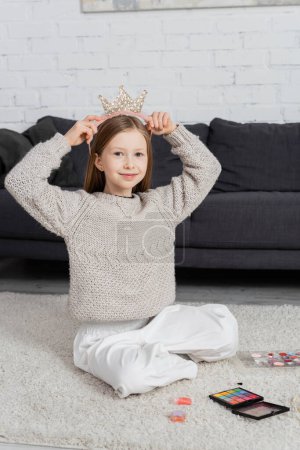 glückliches preteen Mädchen, das Spielzeugkrone justiert, während es auf Teppich in der Nähe der Make-up-Palette sitzt 