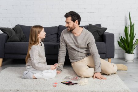 happy father looking at smiling daughter while sitting on carpet near decorative cosmetics 