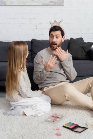 Überraschter Mann mit Spielzeugkrone auf dem Kopf blickt Tochter an, während er neben dekorativer Kosmetik auf Teppich sitzt 