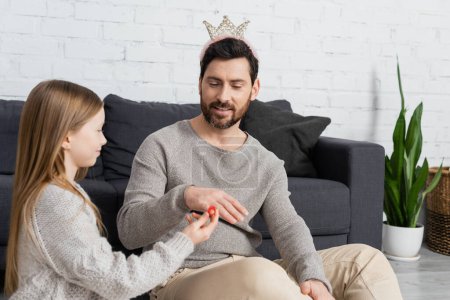 happy girl wearing toy ring on finger of father in crown while playing in living room 