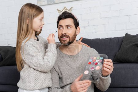 girl holding cosmetic brush while applying makeup on face of smiling father in crown 