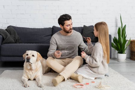 girl holding lip gloss while sitting near father and labrador on carpet 