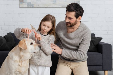glückliches Mädchen mit Spielzeugkrone auf dem Kopf von Labrador neben Vater im Wohnzimmer 