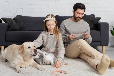 Preteen Mädchen in Spielzeugkrone auf dem Kopf sitzt neben Labrador und Vater beim Schönheitsspiel im Wohnzimmer 