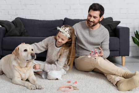 glückliches Mädchen in Spielzeugkrone auf dem Kopf schmusender Labrador neben glücklichem Vater und Make-up-Produkte auf Teppich 