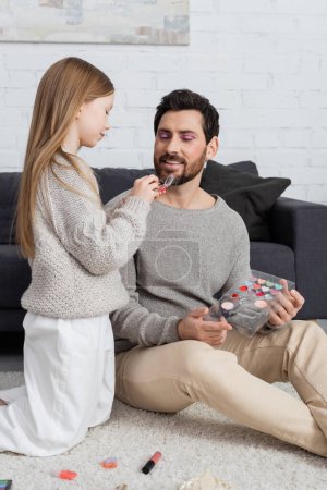 happy father looking at preteen kid holding eye shadow product while playing beauty game 