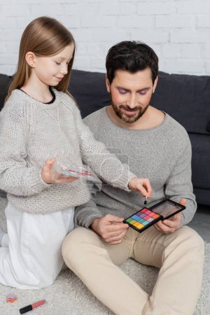 happy father holding eye shadow palette near daughter with cosmetic brush playing beauty game 