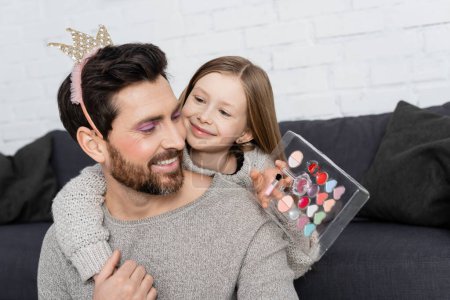 glückliches Kind hält Schönheitspalette und umarmt lächelnden Vater in Spielzeugkrone 