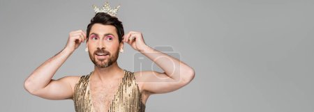 cheerful man in earring wearing toy crown on head while looking away isolated on grey, banner 
