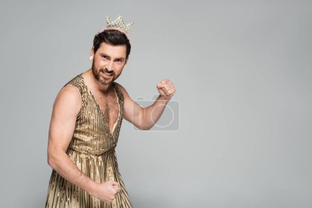 bearded man with toy crown on head and princess dress costume showing muscle isolated on grey 