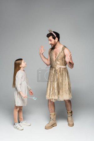 funny father in dress and crown gesturing while looking at daughter with toy wand on grey 