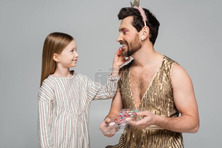 happy girl applying lip gloss on lips of smiling father in dress and crown isolated on grey 