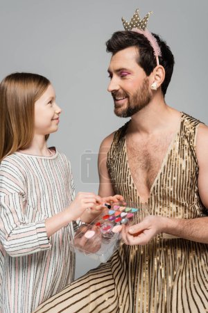 glückliches Kind mit Kosmetikpinsel in der Nähe der Lidschattenpalette und bärtiger Vater in Krone und Kleid isoliert auf grau 