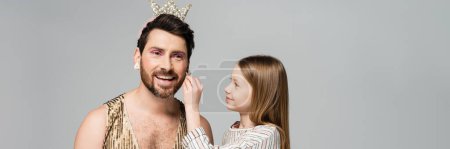 happy kid applying decorative cosmetics on face of bearded father in crown and dress isolated on grey, banner 