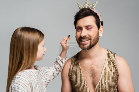kid applying decorative cosmetics on face of happy father in crown isolated on grey
