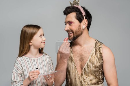 cheerful father in crown applying lip gloss and looking at happy daughter while playing beauty game isolated on grey 