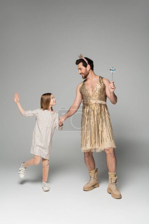 full length of girl in dress holding hands with dad in dress and toy wand on grey 