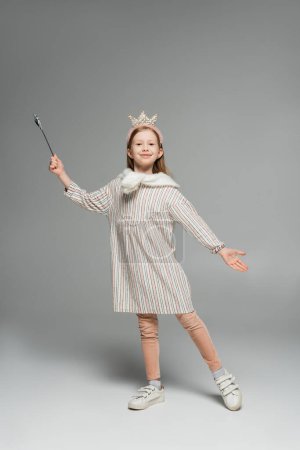 longitud completa de chica feliz en vestido y corona sosteniendo varita de juguete y sonriendo sobre fondo gris 