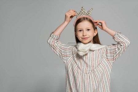happy girl in dress adjusting crown on head and smiling isolated on grey 