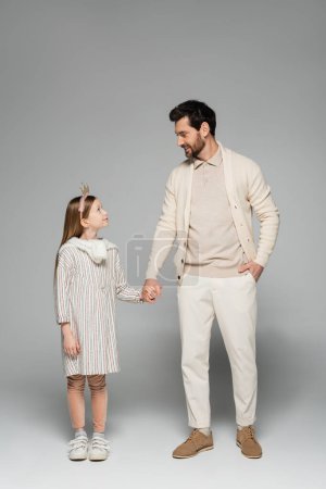 full length of happy man holding hands of kid in crown and standing with hand in pocket on grey