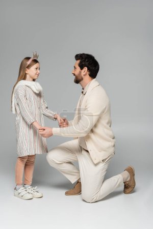 volle Länge eines glücklichen Mannes, der auf Knien steht und die Hände seiner Tochter in Kleid und Krone auf grau hält 