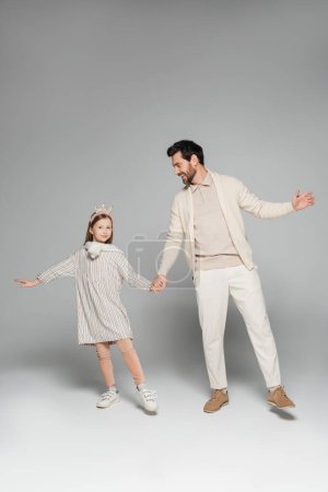 full length of cheerful father and daughter in dress holding hands on grey 