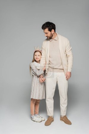full length of happy girl in dress and crown hugging father in white outfit on grey 