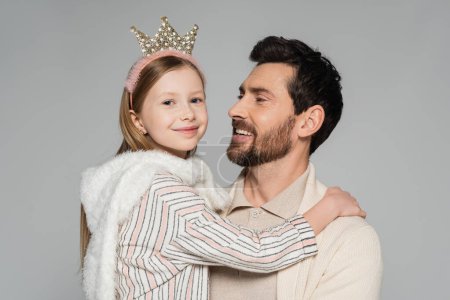 cheerful kid in crown looking at camera while hugging father isolated on grey 
